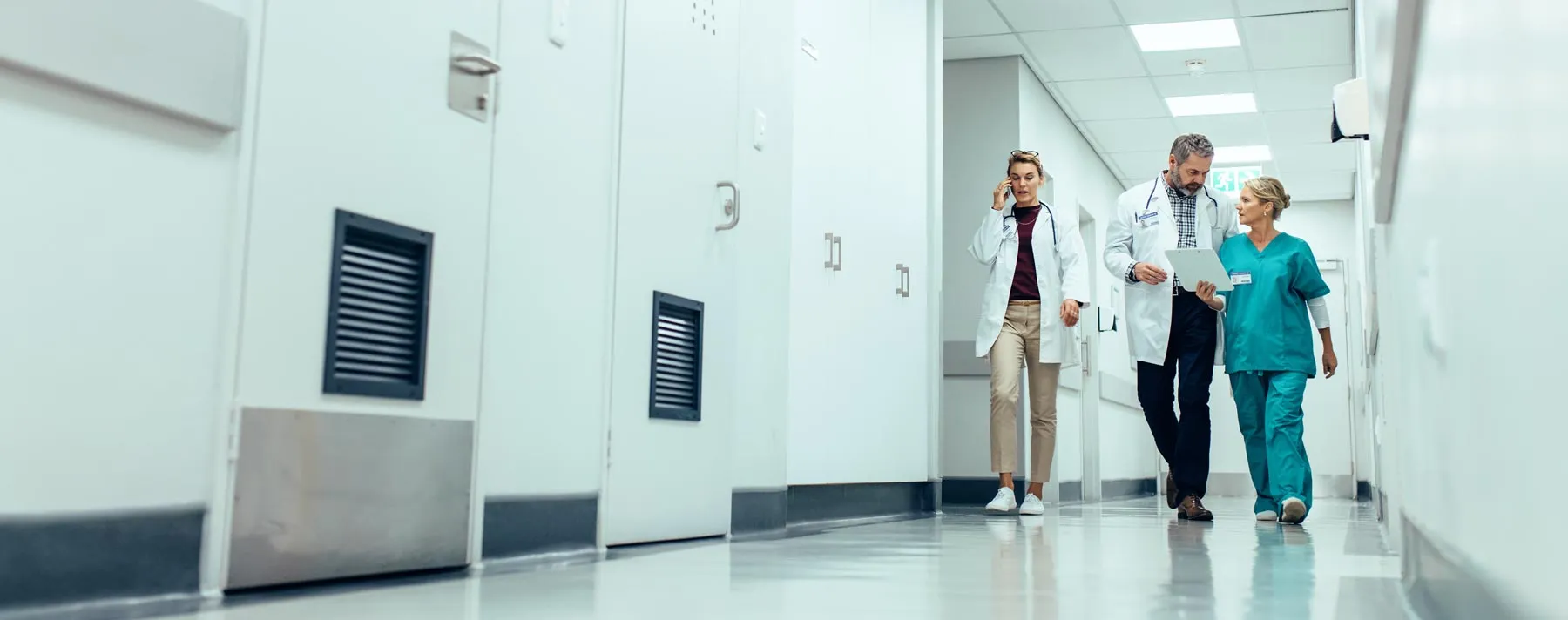 Two doctors and a nurse walking down a hospital corridor, discussing and using a tablet.