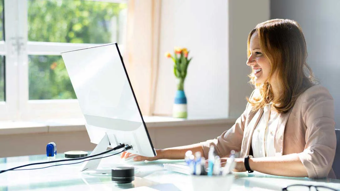 Smiling woman using desktop computer at bright desk near window with vase of flowers.