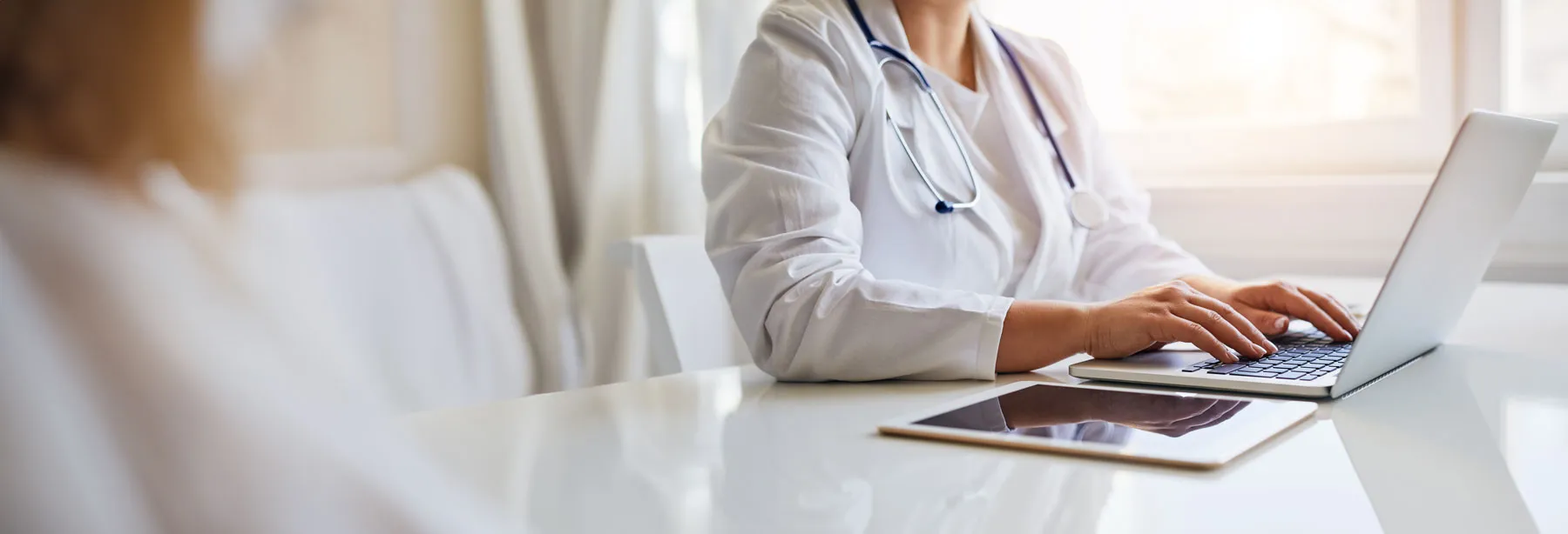 Medical professional in white coat typing on laptop beside tablet on a white desk in bright room.