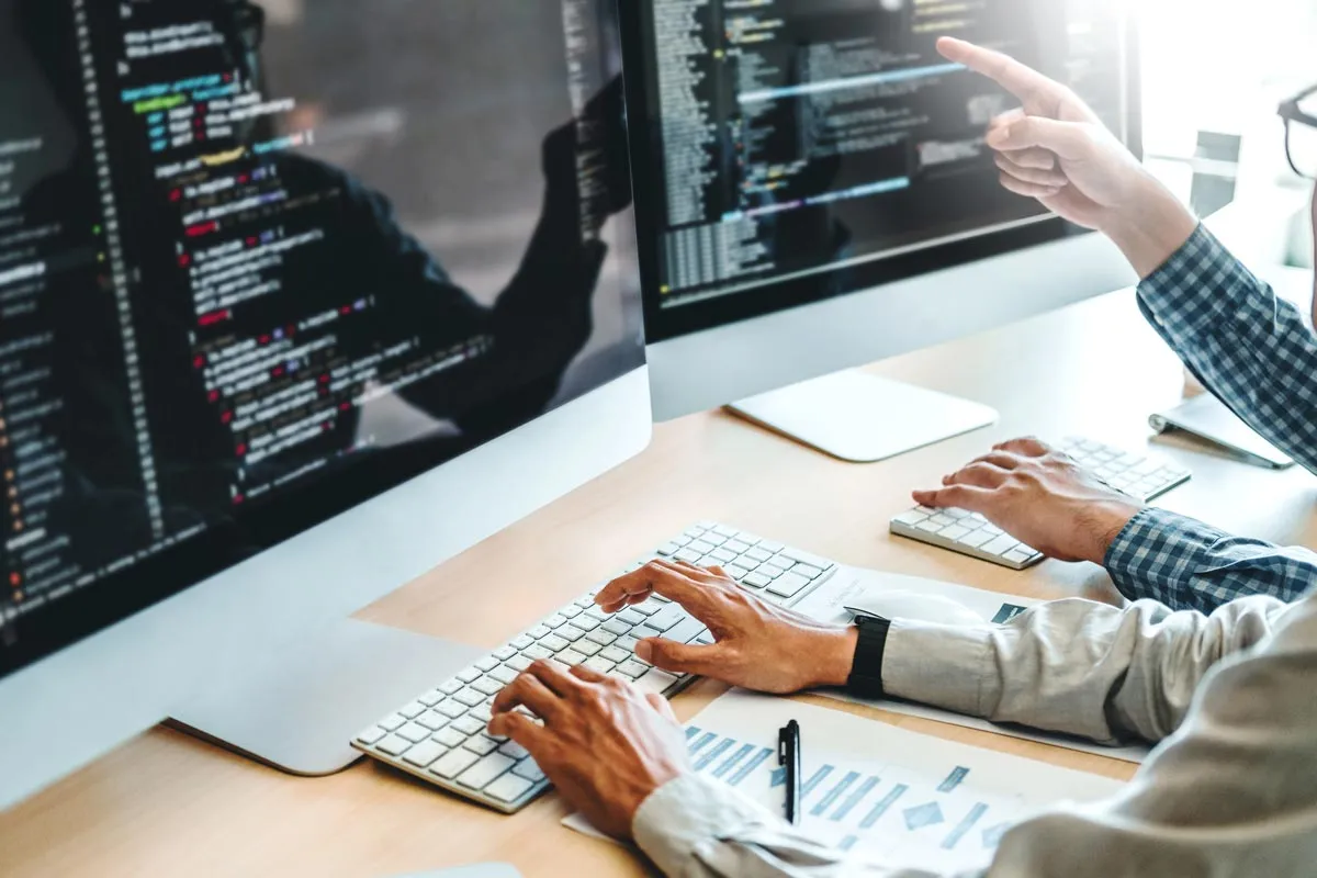 Two people programming on computers with code displayed while one points at a screen in a modern office.
