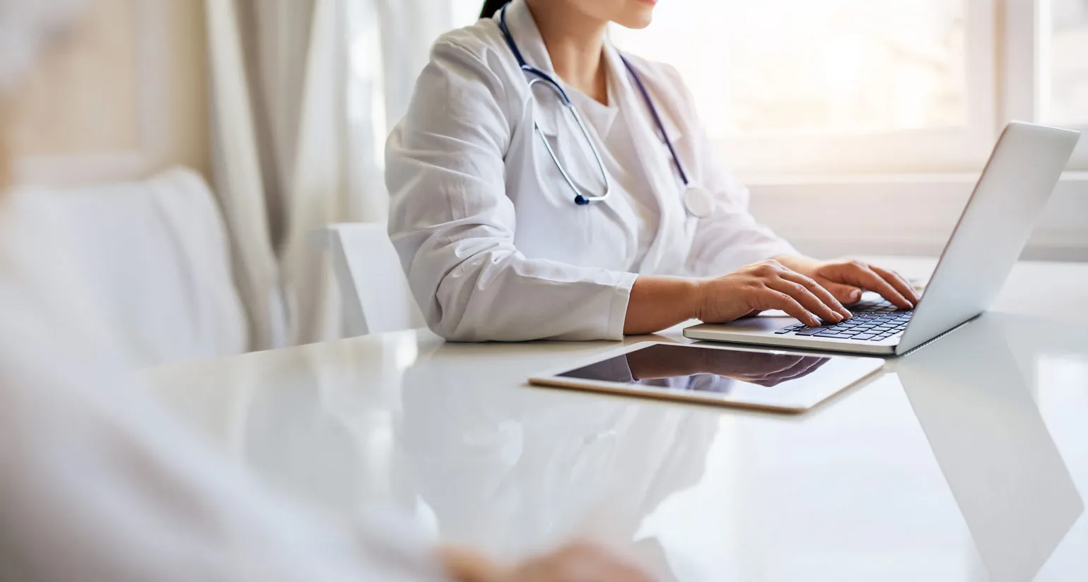 Healthcare professional in white coat with stethoscope using laptop at a white desk with a tablet nearby.