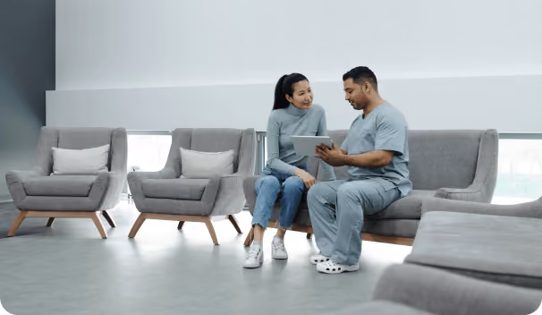 A healthcare professional in scrubs sitting on a gray couch shows a tablet to a woman in a light blue sweater and jeans, presumably discussing medical information in a modern waiting room with gray chairs and large windows.