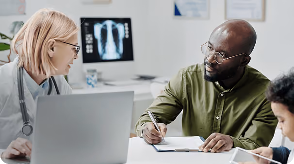 A female doctor with glasses and a stethoscope around her neck consults a male patient who is writing on a clipboard in a medical office with an X-ray image displayed on a screen in the background.