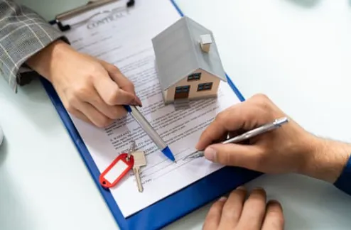 A close-up of two hands signing a contract with a model house and keys on the table.