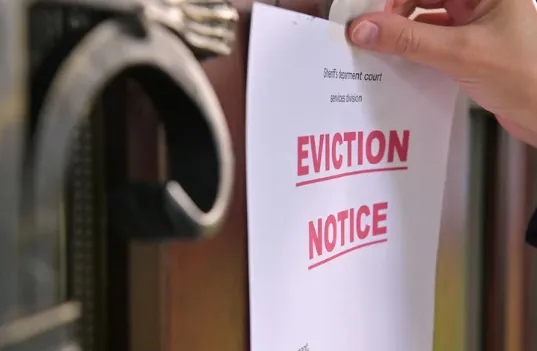 Hand taping an eviction notice to a wooden front door, with bold red lettering.