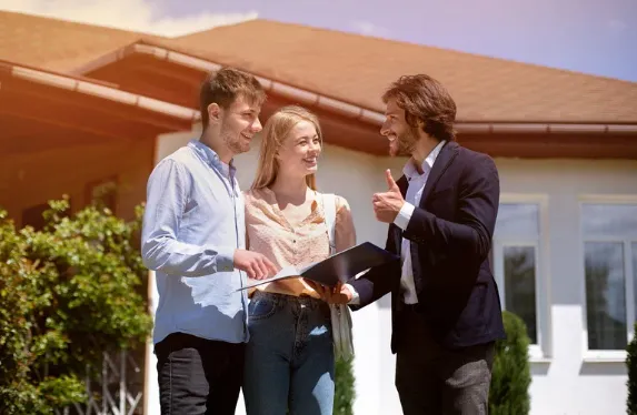 Property manager discussing property options with a happy young couple outside a house—depicting home buying guidance and real estate consultation.