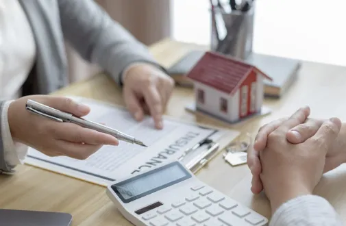 Close-up of two people reviewing insurance documents with a miniature house, calculator, and keys on the table—representing property insurance planning and financial discussions.