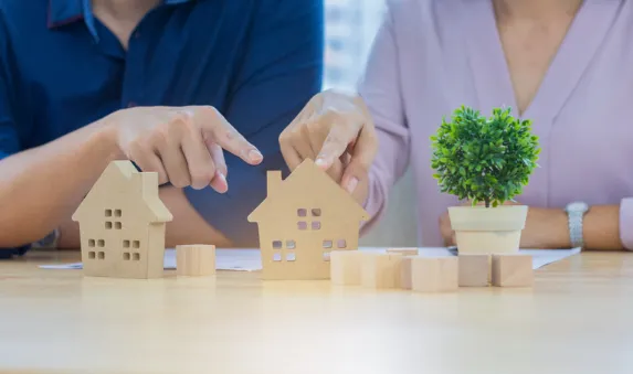 Two people comparing wooden house models on a desk, symbolising property selection, investment planning, or real estate decision-making.
