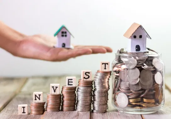 Stacks of coins with wooden blocks spelling "INVEST", a hand holding a miniature house, and another small house sitting on a jar full of coins, symbolising property investment growth.