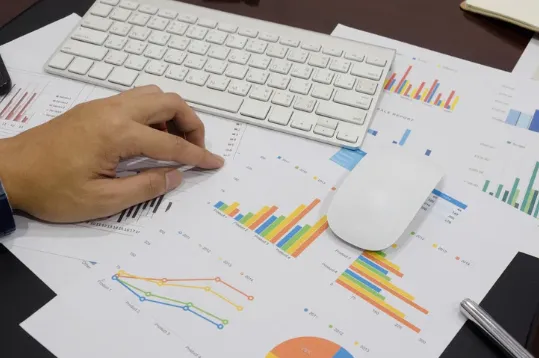 Person analysing colourful financial graphs on printed sheets with a white computer keyboard and mouse, representing investment strategy and data analysis.