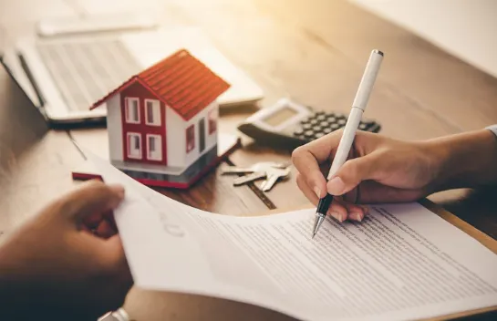 Person signing real estate contract with a model house, calculator, and keys in the background, representing a property purchase or investment agreement