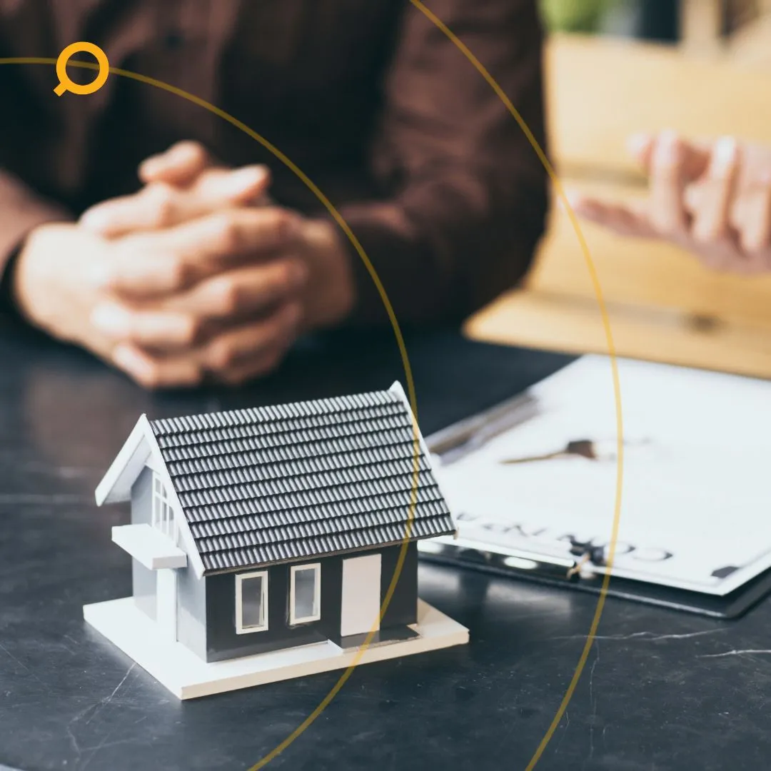 Close-up of a small model house on a desk, with documents and hands gesturing in the background—symbolising real estate investment discussions or planning.