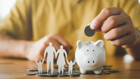 Person placing coin into white piggy bank with paper cut-out family in foreground—symbolising family savings, budgeting, and financial planning.