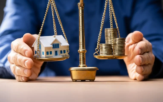 Person balancing a scale with a small house model on one side and stacks of coins on the other, representing real estate value and financial balance.