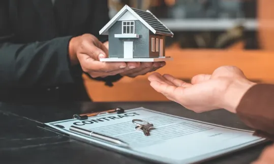 A person hands over a miniature house model to another individual, symbolising a property sale, with a signed contract and house key on the table in the foreground.