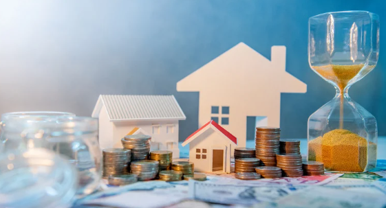 Paper house models with stacked coins, banknotes, glass jars, and an hourglass symbolising real estate investment, savings, and financial planning over time