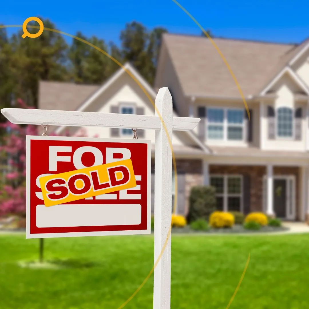 A red "For Sale" sign with a yellow "SOLD" sticker in front of a modern suburban house with a well-kept lawn and flower beds.