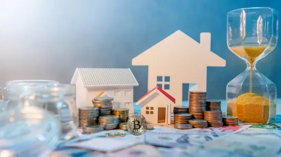Miniature houses surrounded by stacked coins, currency notes, and an hourglass, highlighting the link between property investment, time, and cryptocurrency like Bitcoin.