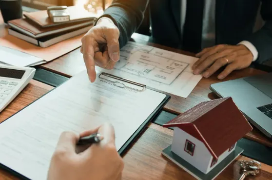 Miniature wooden house, keys, and documents placed on a desk, with two people discussing property investment or home purchase in the background.