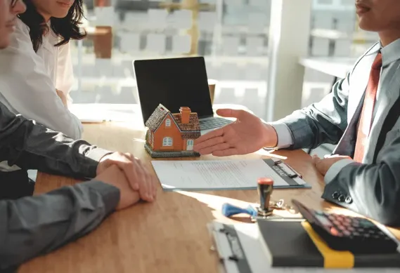 Real estate agent presenting a miniature house to clients during a property purchase meeting, with contract and calculator on the table.