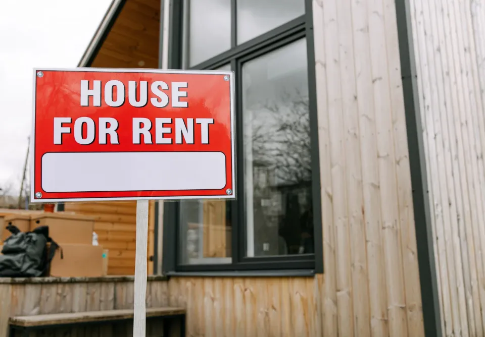 Red and white "House for Rent" sign in front of a modern wooden house with large windows.