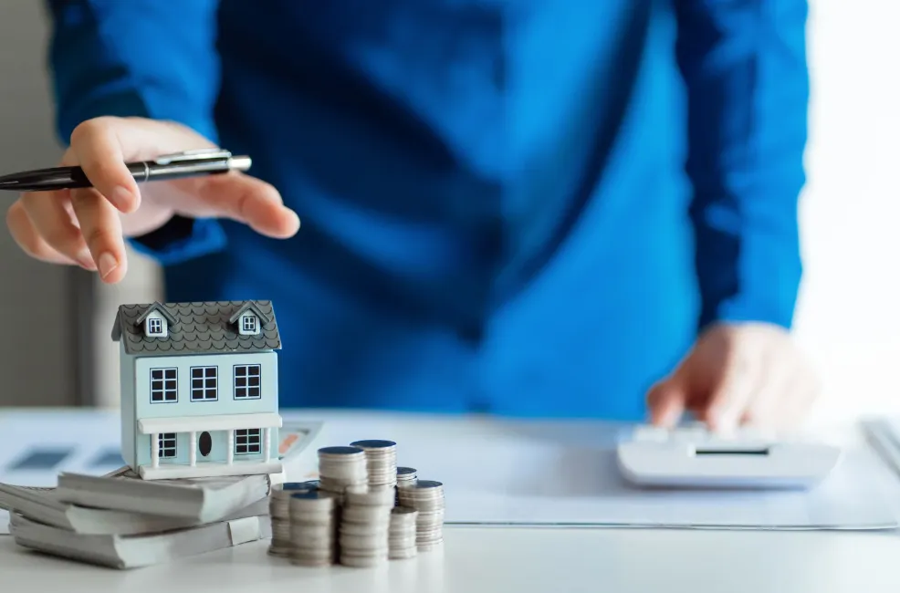 Man in blue suit with pen in hand, reaching for a miniature house placed on top of money bills with coins beside it, alongside a calculator on the table, representing real estate investment and financial planning.