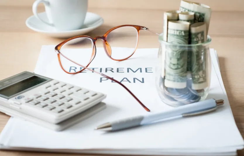 Retirement plan concept with calculator, glasses, rolled dollar bills in a jar, and a pen on financial planning documents.