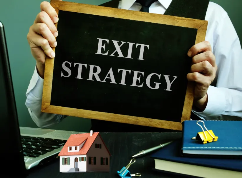 Real estate exit strategy concept with businessman holding a chalkboard and house model on desk.