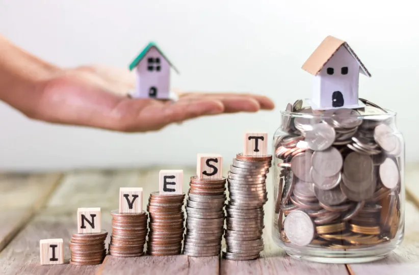 Property investment concept with stacked coins spelling INVEST, miniature houses on a hand and a jar filled with coins.