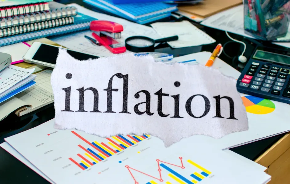 Inflation text with office desk items including calculators, property reports, graphs, notebook, stapler, pencil, magnifying glass, and smartphone symbolising financial analysis and rising costs.