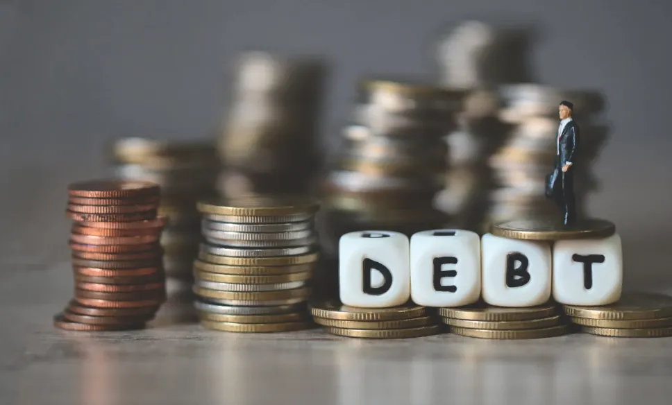 Stacks of coins with dice spelling out DEBT and miniature businessman figure symbolising financial burden and money management.