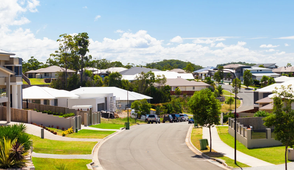 Modern Australian suburban neighbourhood with newly built houses, green lawns, and cars parked along a quiet residential street under a bright sky.