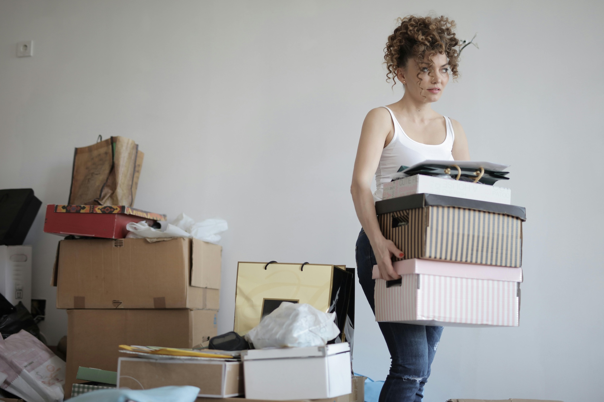 A young woman is carrying multiple boxes, and there are piles of boxes in the background behind her.