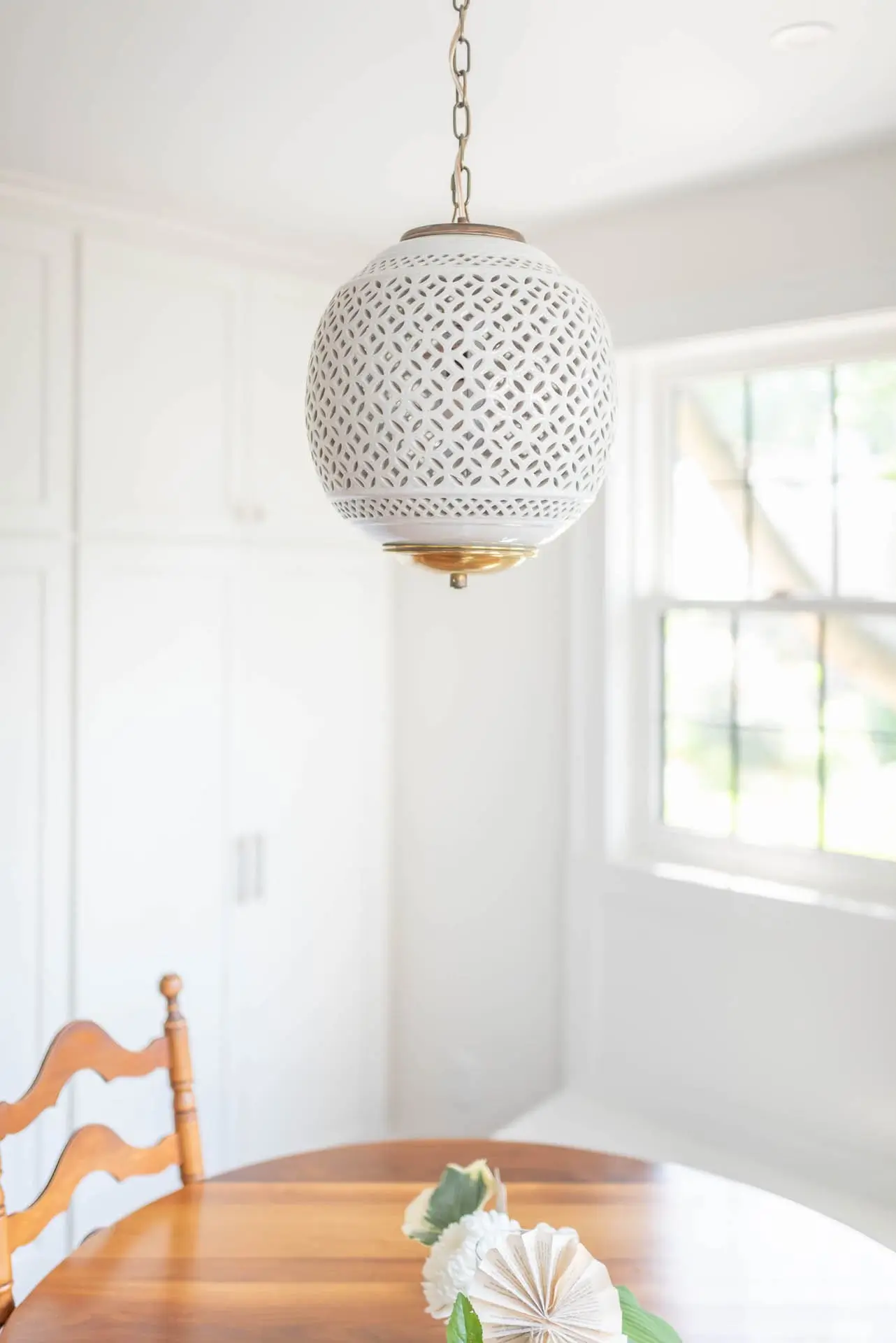 A bright room with white walls featuring a decorative white pendant light hanging above a wooden dining table with a single white flower on it