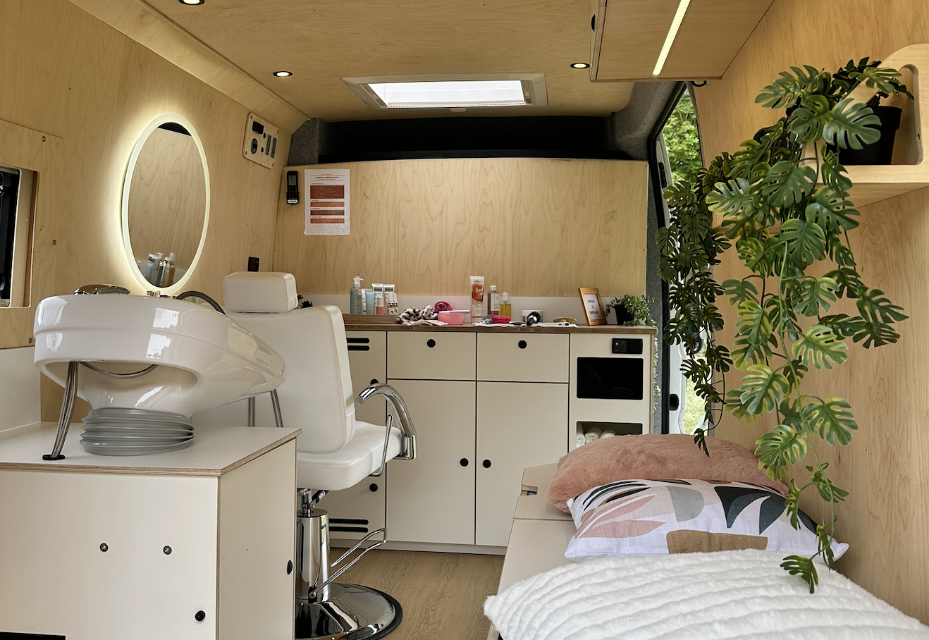 Interior of a small salon with a black styling chair, wash basin, mirror with lights, and wooden surfaces.