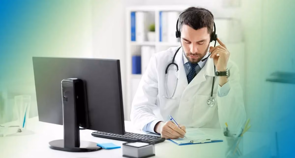 A physician stands at a computer station with a headset helping a patient over the phone.