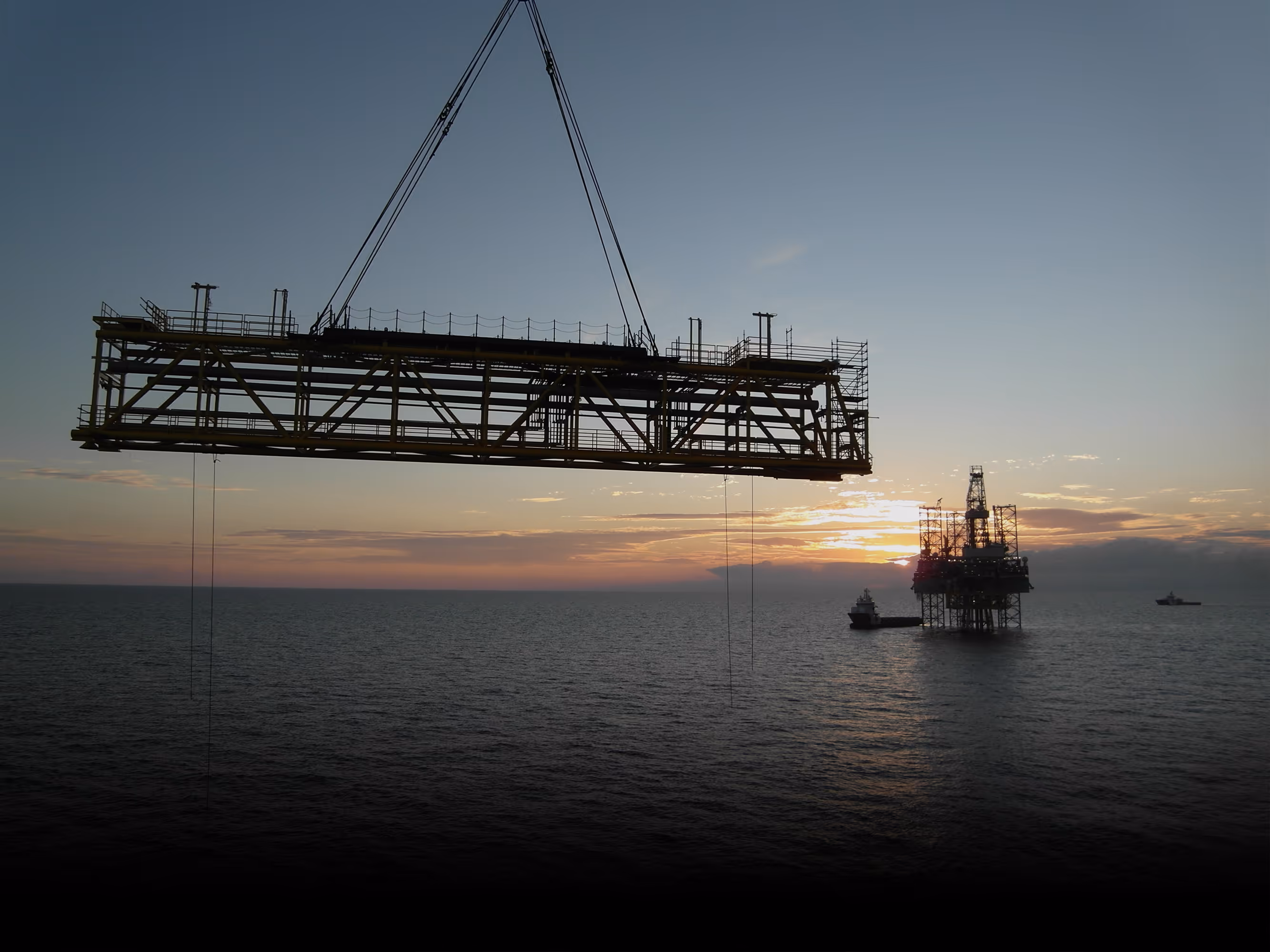 Large metal platform suspended by cables over ocean near offshore oil rig at sunset.