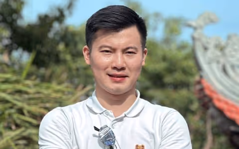 Smiling man in a white polo shirt outdoors with greenery and a traditional architectural structure in the background.