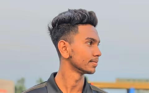 Young man with short styled hair looking to the right against a blurred outdoor background.