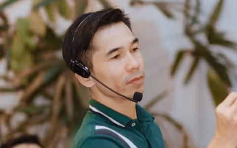 Man wearing a green polo shirt and a headset microphone, looking intently to the side with blurred greenery in the background.