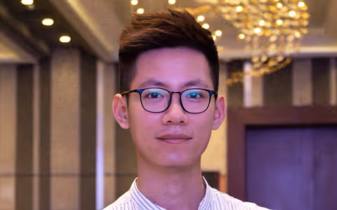 Young man wearing glasses and a striped shirt standing indoors with warm lighting and a chandelier in the background.