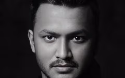 Close-up portrait of a man with dark hair and a mustache against a black background.