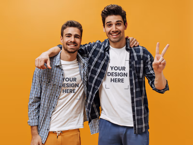 Two smiling men wearing white t-shirts with 'Your design here' text, arms around each other, one making a peace sign, standing against an orange background.