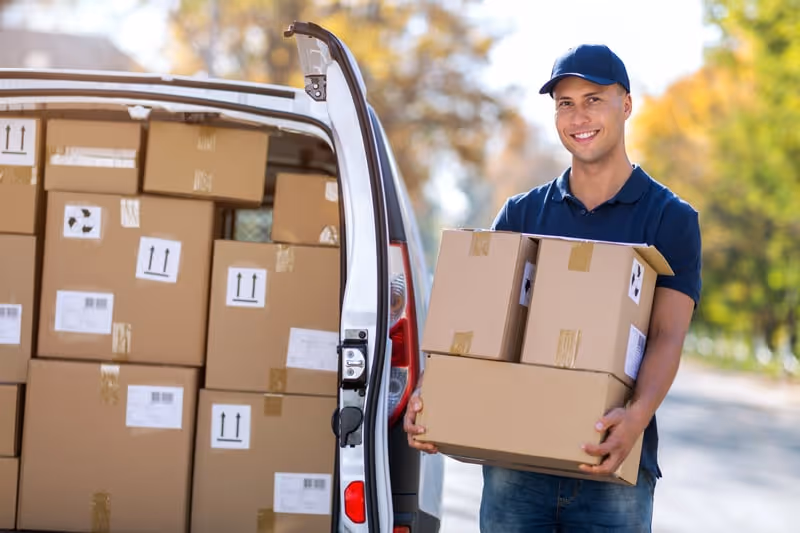 Smiling delivery man wearing a blue cap and shirt holding three cardboard boxes next to an open van filled with more boxes.