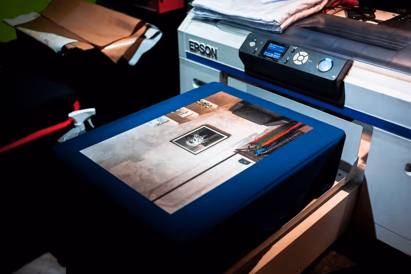An Epson printer printing a photograph with framed art and colorful pens on a table.