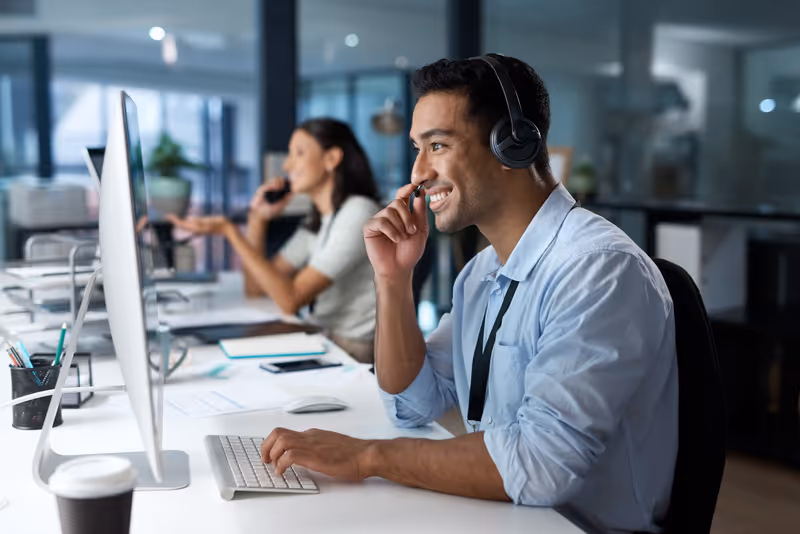Smiling male customer service representative wearing a headset and typing on a keyboard at a desk, with a female colleague on the phone in the background.