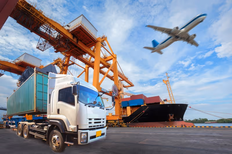 White container truck parked at a port with large orange cranes and a cargo ship loaded with containers, with an airplane flying overhead.