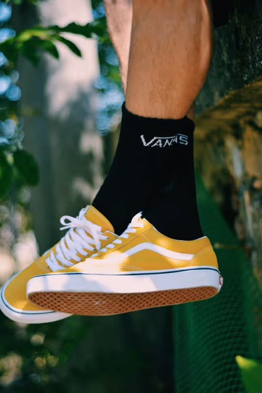 Person wearing yellow Vans sneakers and black Vans socks, legs resting on a ledge outdoors with greenery in the background.