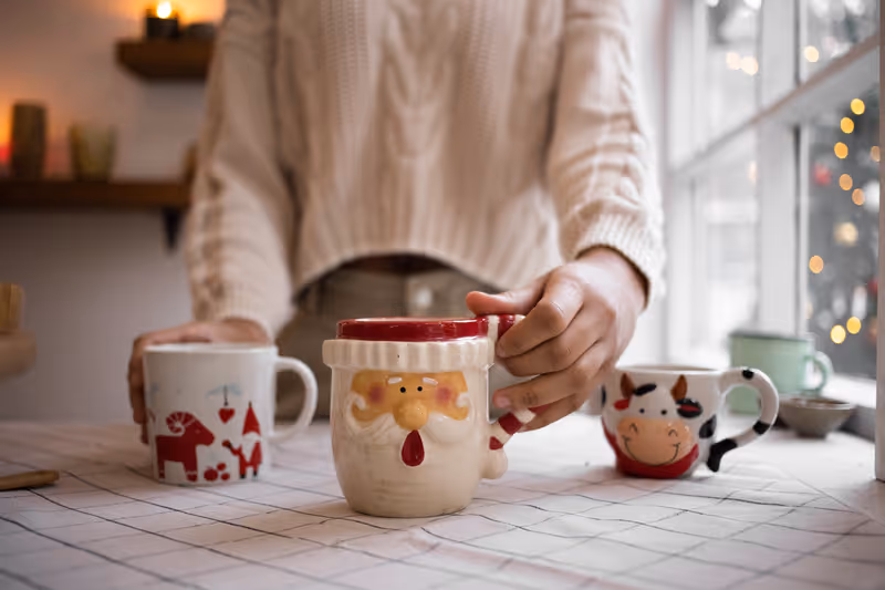 Person holding a festive Santa-themed mug on a table with two other holiday mugs, near a window with blurred Christmas lights.