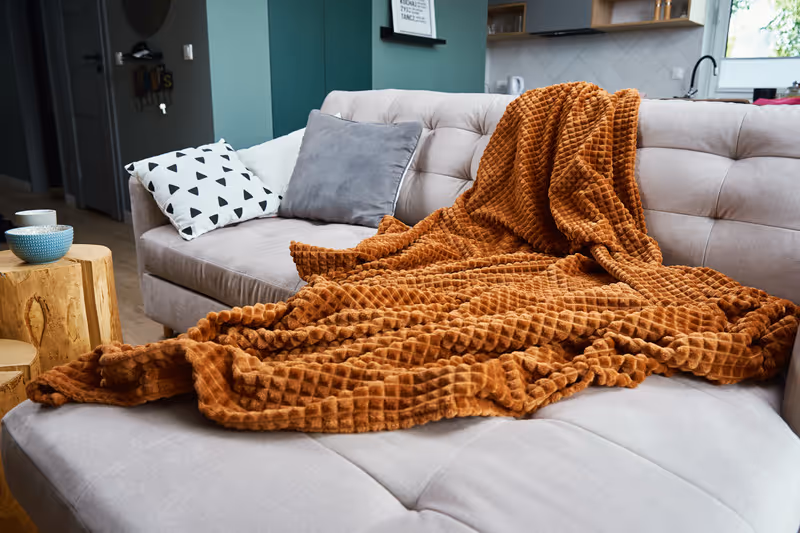 Soft rust-colored textured blanket draped over a beige sofa with gray and patterned pillows in a modern living room.